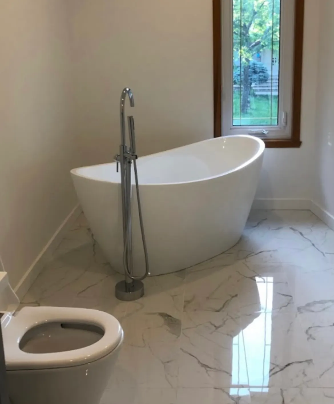 Curved freestanding bathtub with a chrome floor-mounted filler and handshower, on a marble floor.