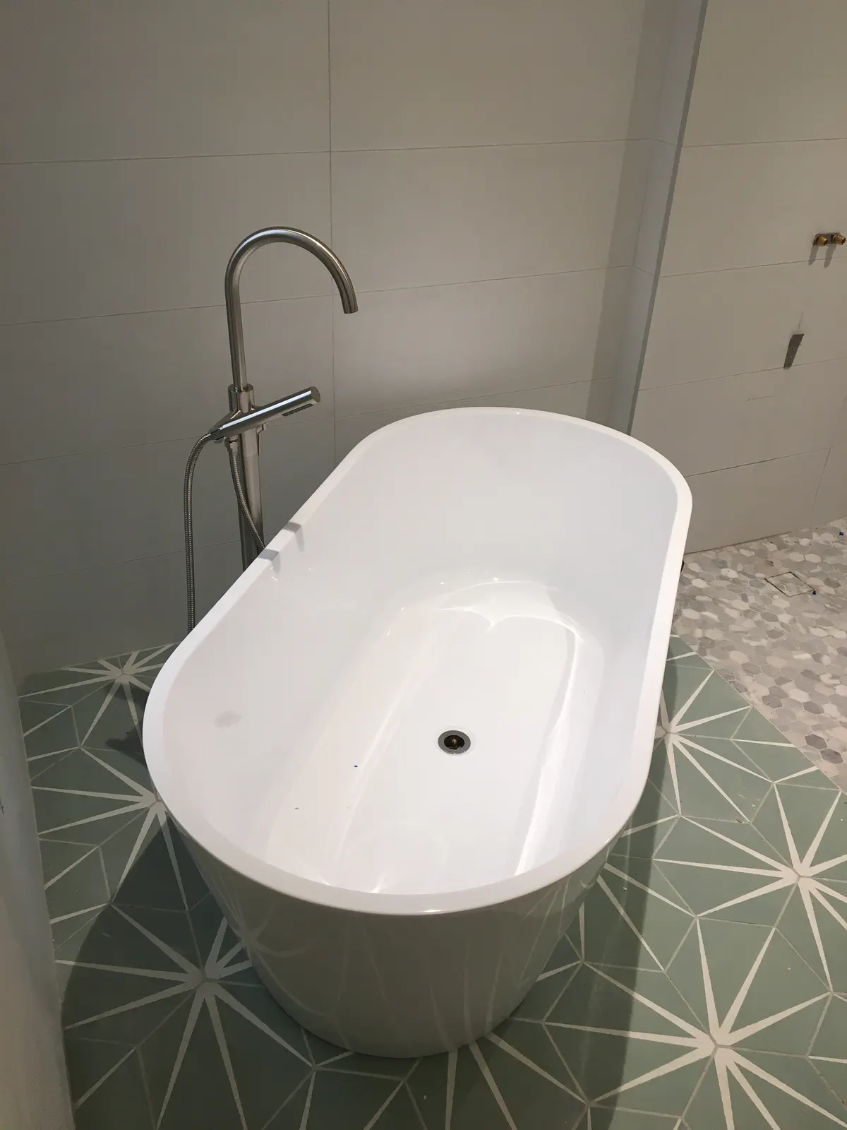 Bain autoportant ovale alimenté par un robinet sur pied, posé sur un plancher de tuiles à motif.