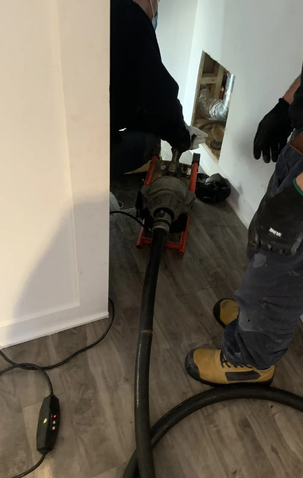 Technicians using a cable machine to clear a drain through an access opening cut in the wall.