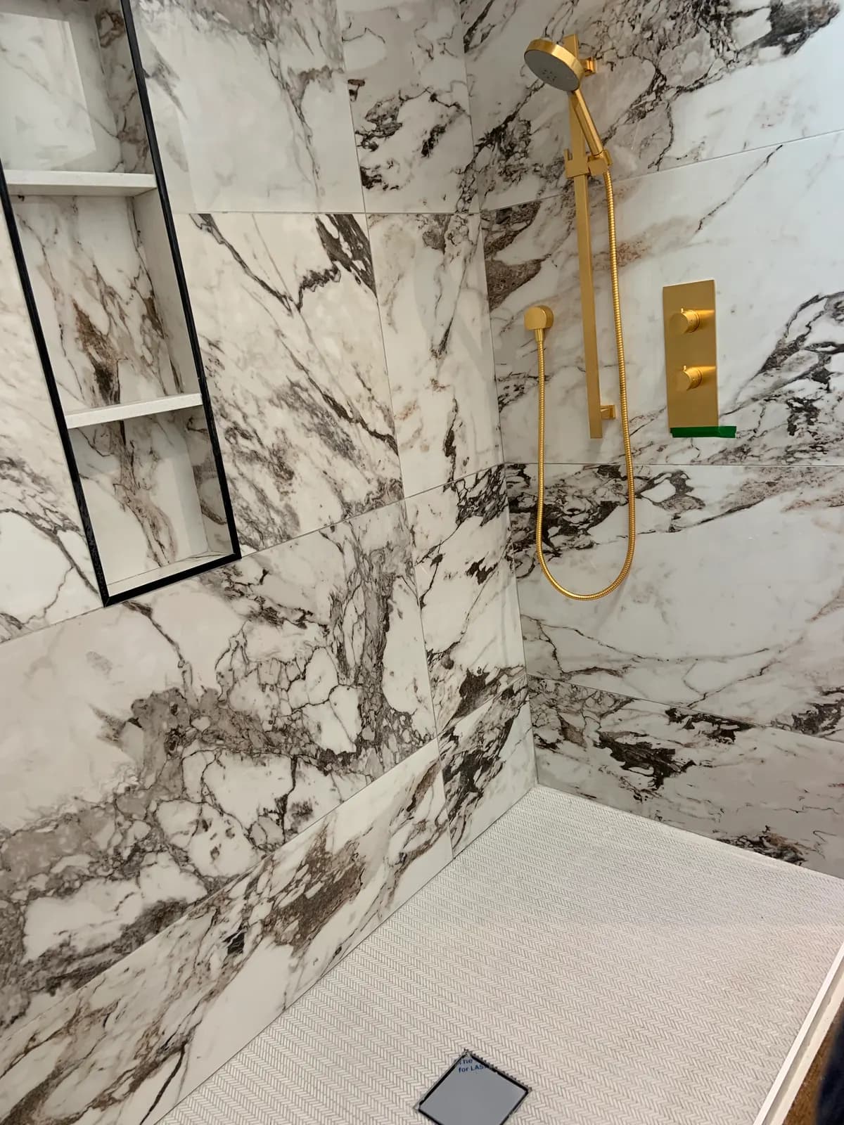 Marble-effect tiled shower with brushed-gold fixtures, a slide-bar handshower, a recessed niche and a square drain.