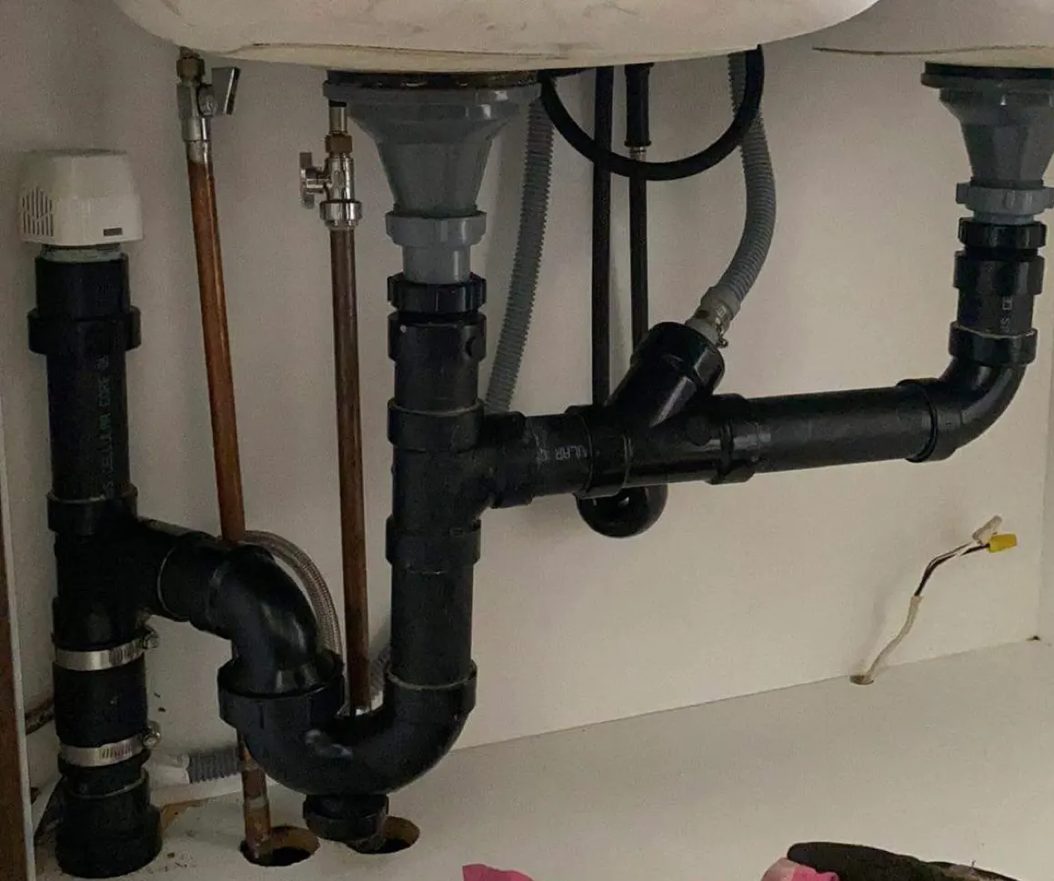 Black ABS drain under a double sink, with an air admittance valve and dishwasher connection.