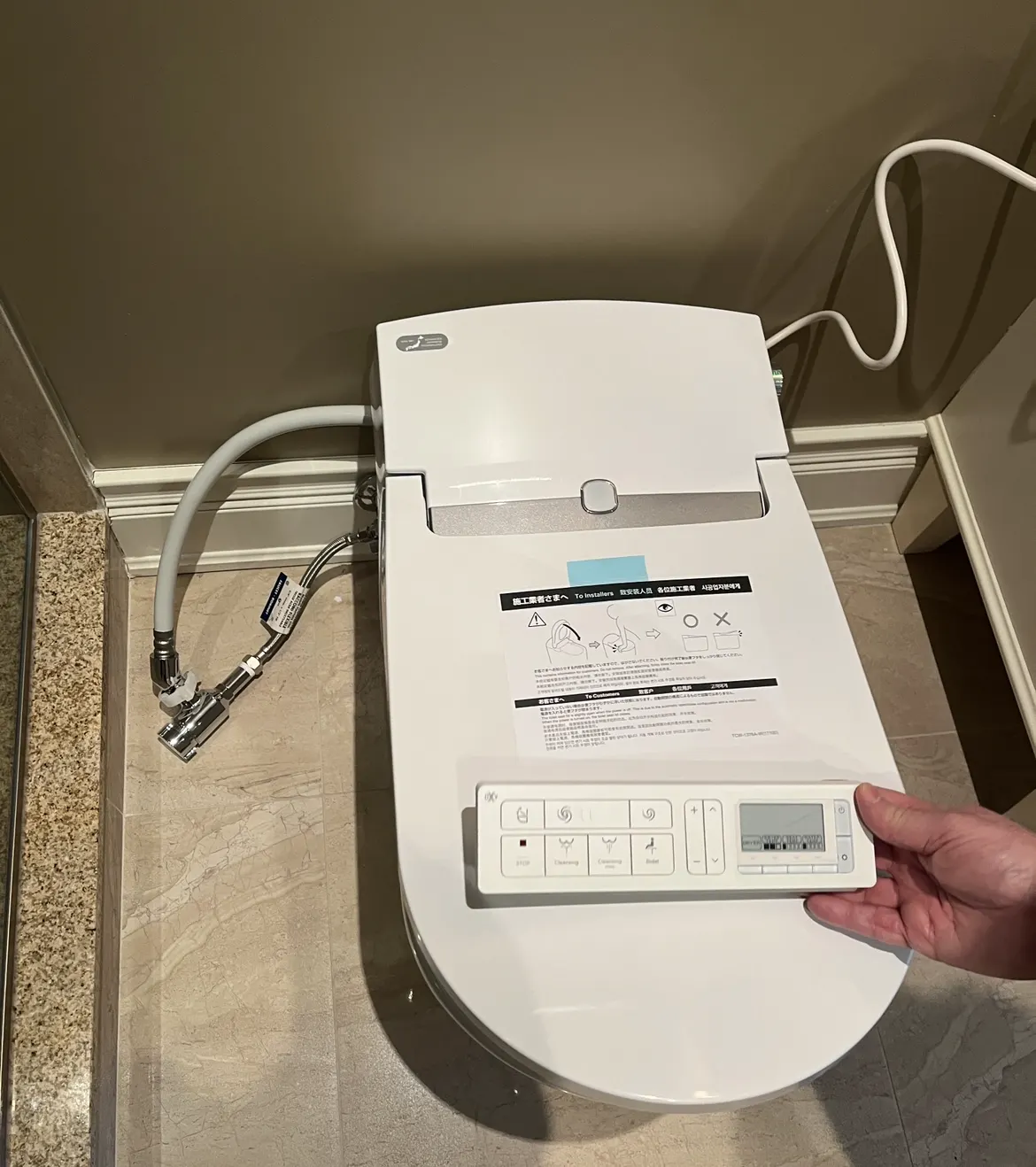 Smart toilet with a bidet seat and remote control, connected to the water supply through a tee fitting.
