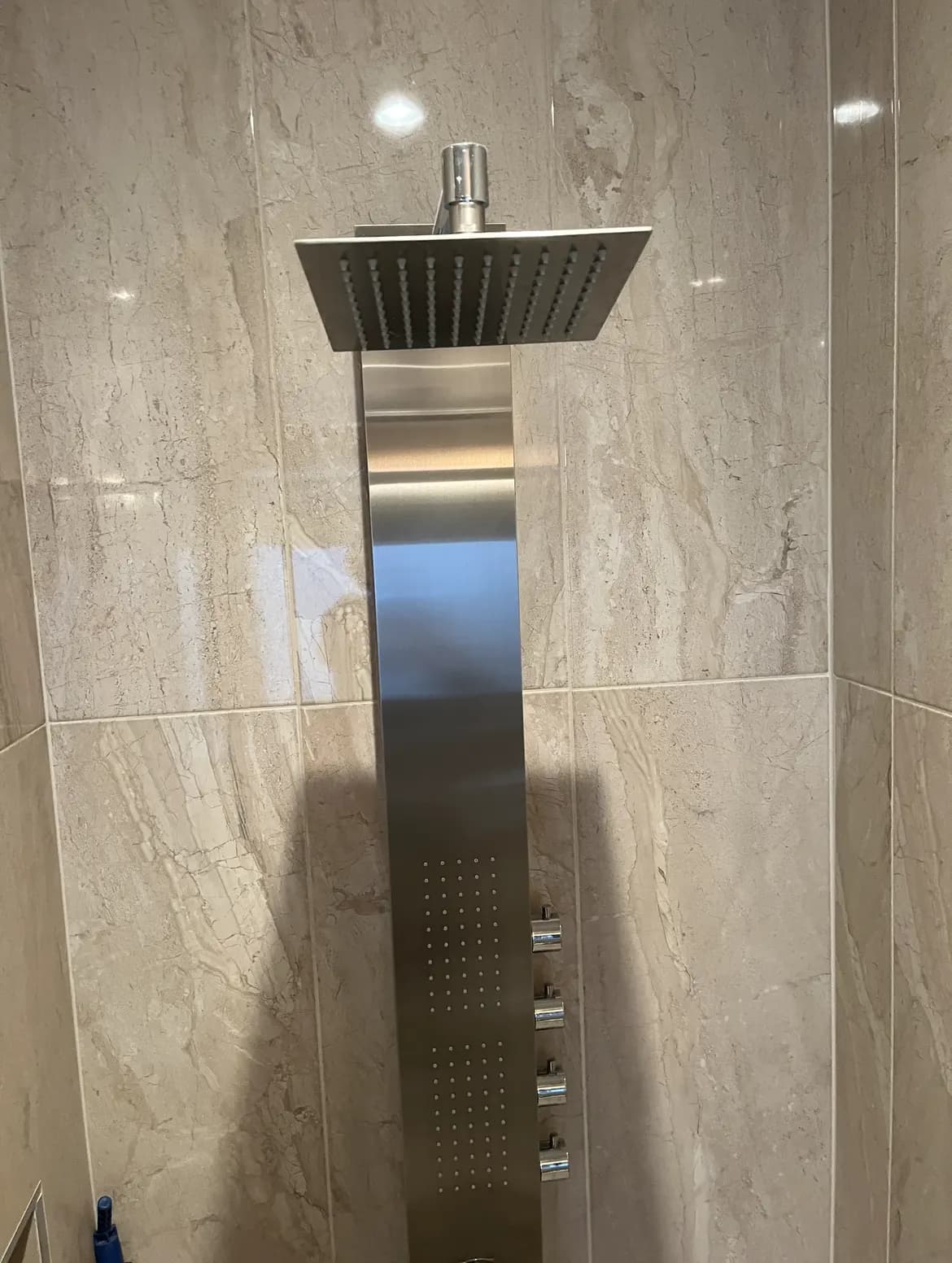 Stainless-steel shower panel with a square rain head and massage jets, on veined beige tile.