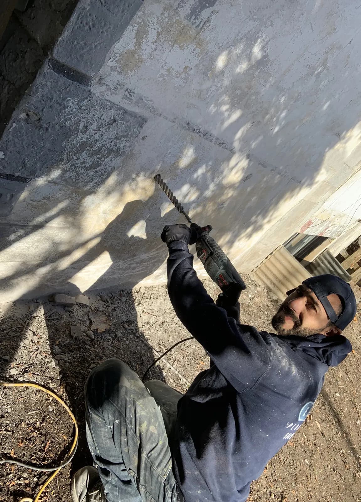 Plumber drilling through a foundation wall with a rotary hammer during outdoor work.