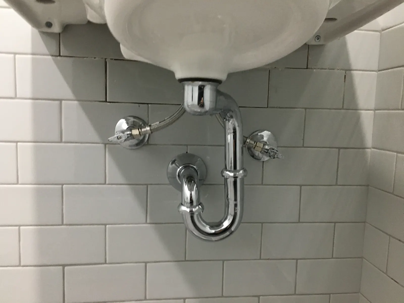 Chrome P-trap and shut-off valves under a wall-mounted basin, against white tile.