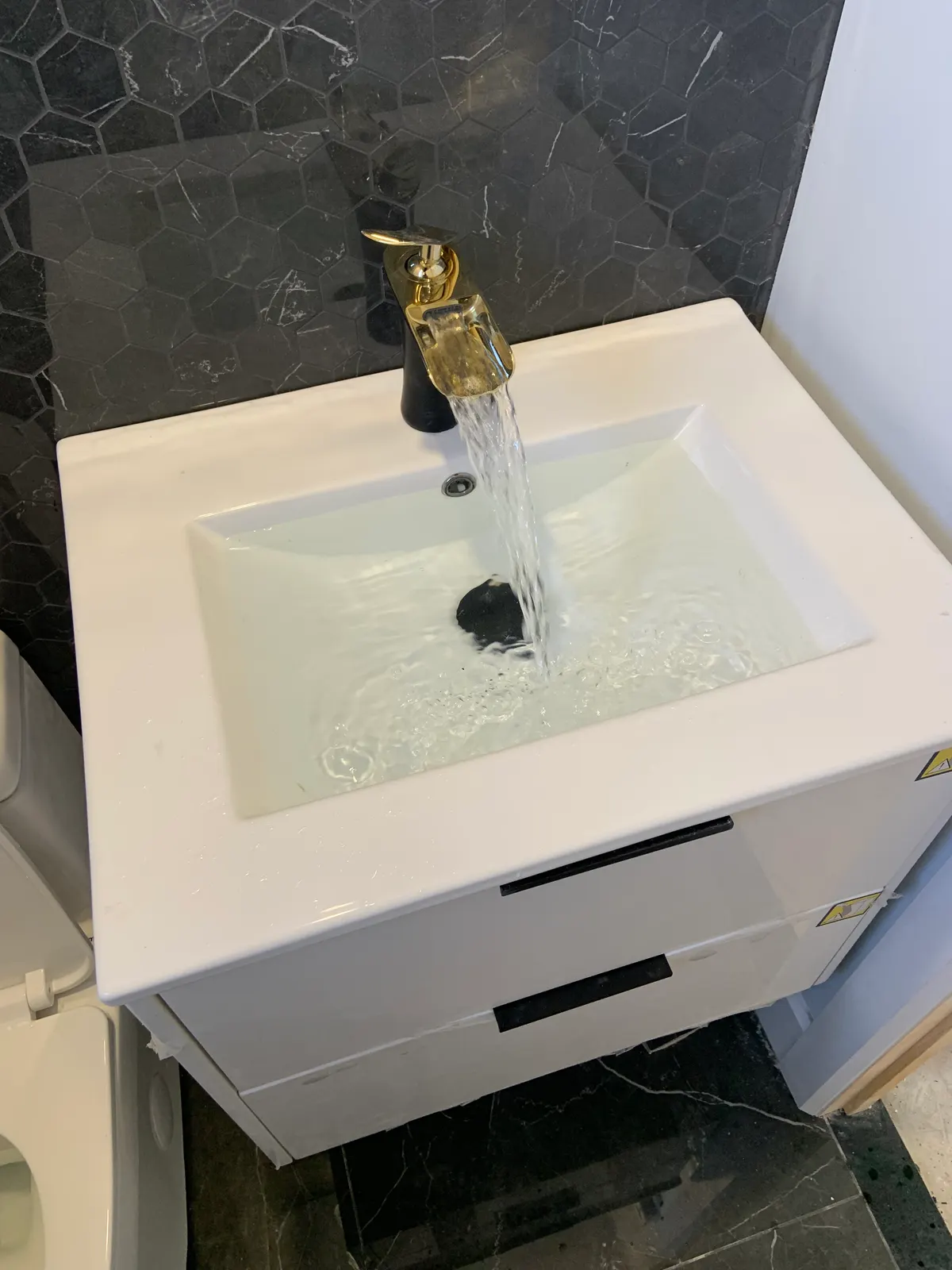White vanity with a running black-and-gold waterfall faucet, in front of veined black hexagonal tile.